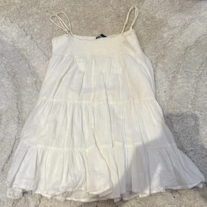 Vit somrig kortklänning från Zara - Supersöt vit kortklänning från Zara med smock upptill och tunna axelband. Klänningen har volanger i flera lager och är tillverkad i ett lätt, luftigt material som passar perfekt för varma dagar. Enkel och stilren design som ger en fräsch look.