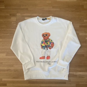 Vit sweatshirt Polo Bear Ralph Lauren - Säljer då den inte används. Vit sweatshirt från Ralph Lauren med ikoniska Polo Bear-trycket framtill. Tröjan har rund hals, långa ärmar och ribbade muddar. Perfekt för dig som gillar streetstyle och vill sticka ut med en klassisk men lekfull look.