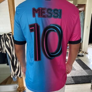 Inter Miami Messi 10 fotbollströja Adidas - Säljer en Inter Miami fotbollströja från Adidas med Messi 10 på ryggen. Tröjan har en snygg färgövergång från blått till rosa, svarta detaljer och klubbmärke på bröstet. Kortärmad modell i lätt och ventilerande material, perfekt för fotbollsplanen.