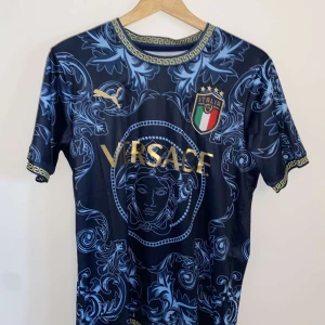 Italien fotbollströja - Snygg marinblå fotbollströja från Puma. Tröjan har guldiga detaljer, Medusa-tryck på bröstet samt Italiens landslagsemblem. Unikt blått mönster över hela tröjan och klassiska grekiska bårder vid hals och ärmslut.