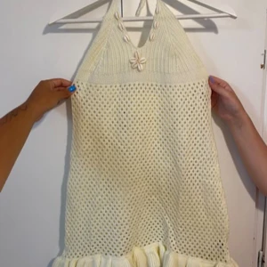 Stickad ljus kortklänning med snäckor - Superfin ljus stickad kortklänning med halterneck och dekorativa snäckor framtill. Klänningen har virkad känsla, volangkant nertill och öppen rygg. Perfekt för sommarens alla äventyr!