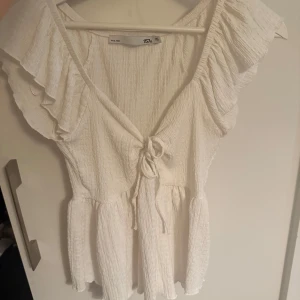 Vit kortklänning med volang och knyt - Supersöt vit blus från 157 med volangärmar och v-ringning. Blusen har knyt framtill och är gjord i ett lätt, krinklat tyg som ger en avslappnad känsla. Perfekt för varma dagar och somriga vibes!