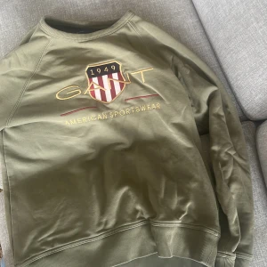 Olivgrön sweatshirt från Gant - Snygg olivgrön sweatshirt från Gant med broderad logga och sköld på bröstet. Tröjan har rund hals, långa ärmar och ribbade muddar. Perfekt för en sportig och avslappnad stil.