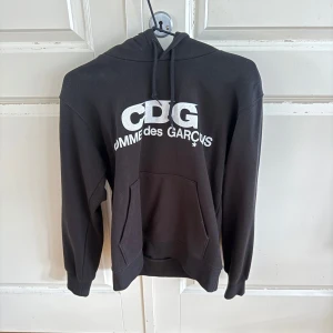 Svart CDG hoodie med tryck - Svart hoodie från Comme des Garçons med stor vit CDG-logga framtill. Klassisk känguruficka, snörning i huvan och mjukt bomullsmaterial. Perfekt för dig som gillar streetwear och vill sticka ut med en ikonisk look.