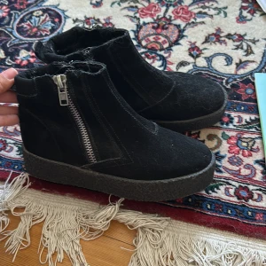 Svarta boots i mocka med dragkedja - Svarta boots i mocka med grov platt sula och dragkedja på sidan. Insidan är fodrad för extra värme och komfort. Snygg rund tå och enkel design som passar till många outfits. Perfekta för kyligare dagar.