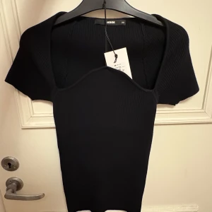 Svart ribbad topp från Bik Bok XS - Supersnygg svart ribbad topp från Bik Bok i figurnära modell. Toppen har korta ärmar och en unik, hjärtformad urringning som ger en cool touch. Perfekt att styla med jeans eller kjol för en trendig look.