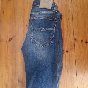 Blå jeans från Nudie Jeans Co - Säljer ett par klassiska blå jeans från Nudie Jeans Co med snygga kontrastsömmar och ikonisk broderad bakficka. Jeansen har normal passform, fem fickor och är tillverkade i slitstark bomull. Perfekta för dig som gillar stilrena och tidlösa jeans.