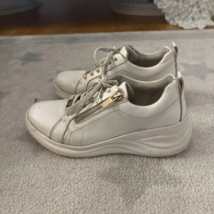 Vita sneakers med gulddetaljer! - Fina vita sneakers med lite högre häl, använda en del men ändå fina. På skorna finns det en dragkedja i guld och även en liten gulddetalj fram! Fina till vardags eller vid nåt festligt sammanhang 