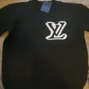 Svart stickad tröja från Louis Vuitton - Svart stickad tröja från Louis Vuitton med stor vit LV-logga framtill. Klassisk rund halsringning och långärmad modell. Perfekt för dig som vill ha en stilren och exklusiv look med streetkänsla.