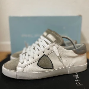 Philippe Model vita sneakers - Snygga vita sneakers från Philippe Model med grå mockadetaljer på tån och tungan. Skorna har en ikonisk sköldlogga på sidan, vita skosnören och en platt sula. Ovandelen är i skinn med kontrasterande grå och orange detaljer vid hälen. Perfekta för en trendig streetstyle. Storlek 40, Bra skick! Box medföljer! Nypris på runt 3000 kr. Hör av er ifall ni har några frågor! MVH Shoeasy ⚜️