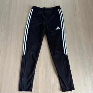 Svarta Adidas träningsbyxor S - Svarta träningsbyxor från Adidas med klassiska vita ränder längs sidorna och logga på låret. Byxorna har smal passform, dragkedja vid benslut och är gjorda i glansig polyester. Perfekta för sport eller chill.