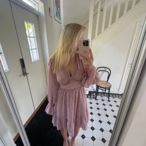 Ljusrosa volangklänning från Dry Lake - Supersöt ljusrosa kortklänning från Dry Lake med v-ringning, volangdetaljer och långa puffärmar. Klänningen har strukturmönster i tyget och smock baktill i midjan. Köpt orginellt för c 1199kr.