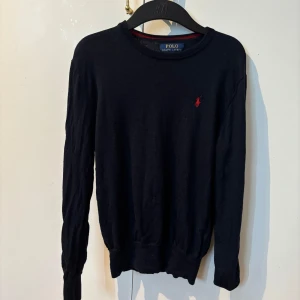 Klassisk svart tröja från Polo Ralph Lauren – 100% bom - Snygg tröja från Polo Ralph Lauren i mörkblått (nästan svart) med det ikoniska röda broderade logot på bröstet. Perfekt för både vardag och finare tillfällen. Tröjan är i ett mjukt och bekvämt bomullsmaterial som andas, med ribbade muddar och fåll för en stilren passform. Det finns inga hål eller fläckar och är i mycket fint skick 😀 passar folk runt 155-160