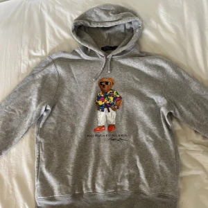 Grå hoodie Polo Bear Ralph Lauren - Hej, säljer en grå hoodie från Polo Ralph Lauren i nyskick med ikoniska Polo Bear-trycket framtill. Tröjan har huva med snörning och känguruficka. Mjuk bomullsblandning och klassisk stil. Snygg detalj med björnen i färgglad skjorta och röda skor.