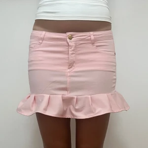 Ljusrosa volangkjol i jeansstil - Supersöt ljusrosa kortkjol med volangkant längst ner. Kjolen har klassisk jeanslook med fickor fram och bak, bälteshällor och knapp framtill. Perfekt för dig som vill ha en feminin och trendig vibe.