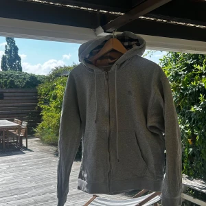 Grå hoodie från Burberry med dragkedja - Säljer en grå hoodie från Burberry med klassiskt rutigt foder i huvan. Tröjan har dragkedja framtill, snörning i huvan och två stora fickor. Perfekt för dig som gillar stilren design med en touch av lyx.