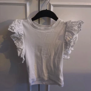 Vit blus med volangärmar - Supersöt vit blus med korta volangärmar i broderad bomull. Ärmarna har ett blommigt hålmönster som ger en romantisk vibe. Perfekt för dig som gillar detaljer och vill sticka ut lite extra. Rak passform och rund halsringning.