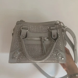 Grå handväska med nitar och dragkedja - Snygg grå handväska med silvriga nitar och detaljer. Väskan har både handtag och axelrem, samt en dragkedja framtill. Perfekt för dig som vill ha en cool och trendig accessoar med lite edge. Helt oanvänd.