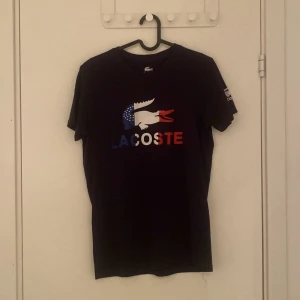 Mörkblå Lacoste t-shirt med tryck - Mörkblå t-shirt från Lacoste med stort färgglatt krokodiltryck och text i blått, vitt och rött på bröstet. Klassisk rund hals och korta ärmar. Lacoste-logga även på ena ärmen. Skön och mjuk bomull, perfekt för en chill och stilren look. Pris kan diskuteras vid snabb affär!