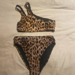 Leopardmönstrad bikini från NA-KD XS - Snygg bikini från NA-KD i leopardmönster med bruna och svarta toner. Toppen har dubbla axelband och en sportig, asymmetrisk form. Bikinin är tillverkad i stretchigt syntetmaterial och är perfekt för stranden eller poolen.