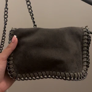 Grå mocka axelväska med kedja - Snygg grå axelväska i mocka med grov kedja som axelrem och coola detaljer längs kanterna. Perfekt storlek för det viktigaste och ger en edgy vibe till din outfit.