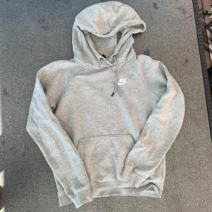 Grå hoodie från Nike med huva - Snygg grå hoodie från Nike i mjuk bomullsblandning. Klassisk modell med huva och snörning, samt stor magficka framtill. Perfekt för chill dagar eller när du vill ha en sportig look. Diskret Nike-logga på bröstet och ribbade muddar vid ärmslut och nederkant.