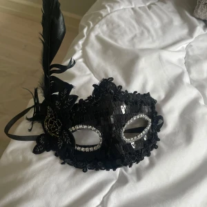 Svart maskeradmask med fjäder - Snygg svart maskeradmask dekorerad med paljetter, glittriga stenar runt ögonen och en stor svart fjäder på sidan. Masken har även en svart blomdetalj med guldbroderi och knyts med band. Perfekt för maskerad eller bal.