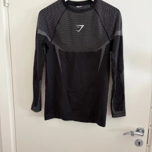 Gymshark Onyx - Svart långärmad träningströja från Gymshark med gråa detaljer och mönstrad mesh på axlar och ärmar. Tight passform som sitter snyggt på kroppen, perfekt för gymmet eller löpning. Snygg sportig look med diskret logga på bröstet.