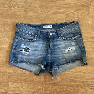 Blå jeansshorts med nitar från Lindex  - Snygga blå jeansshorts från Denim med slitna detaljer, råa kanter och coola silverfärgade nitar på fickorna. Klassisk femficksmodell med låg midja och avslappnad passform. Perfekta för sommaren och ger en edgy vibe till din outfit.