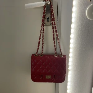 Röd quiltad axelväska med kedja - Snygg röd axelväska i quiltat skinn med guldfärgad kedja och vridlås. Klassisk rektangulär form och elegant design, perfekt för att lyfta din outfit. Väskan har en lång kedjerem som kan bäras både enkel och dubbel.