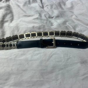 Svart skärp med silverdetaljer Vera Pelle - Unikt svart skärp från Vera Pelle i äkta skinn med massor av blänkande silverdetaljer och glittrande stenar. Bältet har rektangulära och runda dekorationer längs hela bandet och en klassisk silverfärgad spänne. Perfekt för att lyfta vilken outfit som helst.