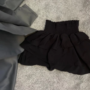 Svart volangkjol från H&M - Supersöt svart kortkjol från H&M med bred smock i midjan och dubbla volanger. Kjolen är luftig och har en riktigt skön passform som sitter snyggt på höften. 