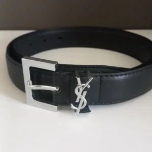 Svart YSL bälte i skinn med silverdetaljer - Snyggt svart bälte från YSL i äkta skinn med en stilren, fyrkantig silverspänne och ikoniska YSL-loggan i metall. Bältet har en klassisk design som lyfter vilken outfit som helst och passar perfekt till jeans eller kostymbyxor.