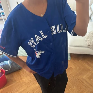 Blå Blue Jays tröja Martin 55 - Säljer en blå Blue Jays supportertröja med tryck fram och bak. På framsidan står det 'Blue Jays' med lagets logga och på ryggen står det 'Martin 55' i vitt. Tröjan har korta ärmar, knappar framtill och är gjord i ett lätt, sportigt material.