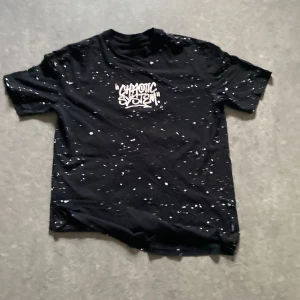Svart t-shirt med graffiti från FSBN - Svart t-shirt från FSBN med relaxed fit och coolt graffiti-tryck både fram och bak. T-shirten har vita stänk över hela tyget och texten 'Chaotic System' i stor stil. Perfekt för dig som gillar streetwear vibes.