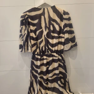 Zebramönstrad kortklänning med vida ärmar - Cool kortklänning med svart och beige zebramönster. Klänningen har vida, korta ärmar och markerad midja med lätt rynk. Luftigt material som ger en avslappnad och trendig vibe. Perfekt för dig som vill sticka ut med ett djurmönster.