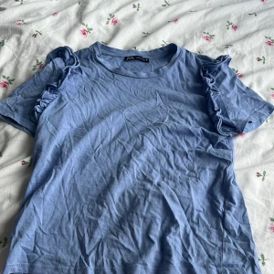 Blå volangtopp från Zara - Supersöt ljusblå topp från Zara med korta ärmar och volangdetaljer på axlarna. T-shirten är i mjuk bomull.