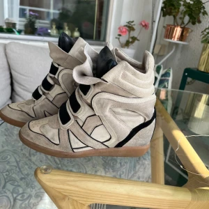  Isabel Marant skor  - Snygga sneakers från Isabel Marant i beige mocka med svarta detaljer och kardborreband. Skorna har en dold kilklack, rund tå och gummisula. Perfekt för dig som vill ha en cool och sportig look med extra höjd.