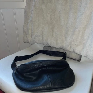 Svart magväska i skinn från Puccini - Snygg svart magväska från Puccini i äkta skinn. Väskan har en justerbar rem med spänne och dragkedja upptill. Perfekt för att bära det viktigaste och ger en clean look till din outfit.