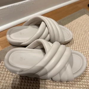 Beiga vadderade sandaler från H&M - Säljer ett par trendiga beiga sandaler från H&M med breda, vadderade korslagda remmar. De har en chunky look och platt sula, perfekta för chill dagar. Tillverkade i syntetmaterial och riktigt sköna att glida i.