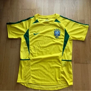 Brasilien fotbollströja Nike L - Gul Brasilien fotbollströja från Nike i storlek L. Tröjan har gröna detaljer längs sidorna och axlarna, rund hals och korta ärmar. Tillverkad i lätt och ventilerande polyester, perfekt för match eller träning. Klassisk landslagsdesign med broderad Brasilien-logga på bröstet.