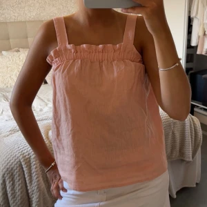 Ljusrosa linnetopp med volangkant - Supersöt ljusrosa linnetopp med breda axelband och volangdetalj upptill. Luftig och perfekt för varma dagar, passar snyggt till både jeans och kjol. Enkel och stilren design som ger en fräsch look.