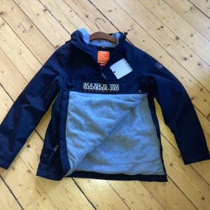 Marinblå jacka från Napapijri M - Snygg marinblå vindjacka från Napapijri med stor logga framtill. Jackan har huva, dragkedja i sidan och grått mjukt foder. Perfekt för blåsiga dagar och har en sportig vibe. Modellen heter Rainforest och är riktigt populär bland ungdomar.