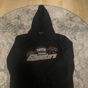 Svart Trapstar hoodie London Shooters - Svart hoodie från Trapstar med stor broderad London Shooters-logga och pantermotiv på bröstet. Klassisk huva med metallöljetter och relaxed fit. Perfekt för dig som gillar streetwear och vill sticka ut med en fet logga. Använd och har inte snören.