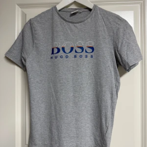 Grå t-shirt från Hugo Boss - Snygg grå t-shirt från Hugo Boss med klassisk rund hals och tryckt logga i blått, vitt och rött framtill. Skön passform och tillverkad i mjuk bomull. Perfekt för en clean och stilren look.