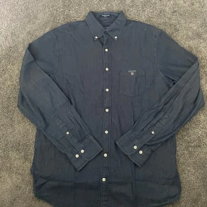 Mörkblå skjorta från GANT  - Snygg mörkblå skjorta från GANT i bomull med klassisk krage och knäppning framtill. Skjortan har en bröstficka med broderad GANT-logga och vita knappar. Passformen är normal och materialet känns mjukt och bekvämt. Skriv vid minsta lilla fundering