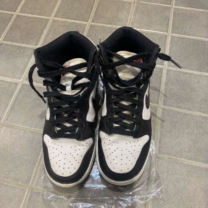 Nike Dunk High svart/vit sneakers - Säljer ett par Nike Dunk High sneakers i svart och vitt. Klassisk siluett med svart Swoosh, perforerad tå och snörning. Ovandel i skinn och textilfoder för skön passform. Perfekta för dig som gillar streetstyle och vill sticka ut med en ikonisk modell.