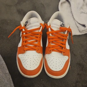 Nike Dunk Low orange och vita sneakers - Nike Dunk Low sneakers i vitt och orange med svarta swooshes dekorerade med stjärnmönster. Skorna har orange snörning, perforerad tå och klassisk låg siluett. Ovandelen är i skinn och de har en platt sula. Perfekta för dig som vill sticka ut med en unik färgkombo.