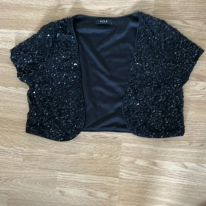 Svart paljett-bolerotopp från Vila - Glittrig svart bolerotopp från Vila, täckt med svarta paljetter som ger en riktigt festlig vibe. Kortärmad och croppad modell med öppen front, perfekt att slänga över en klänning eller topp för extra bling. Snygg och enkel att styla till olika looks.