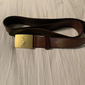 Brunt skärp från Polo Ralph Lauren - Snyggt brunt bälte i skinn från Polo Ralph Lauren med en rejäl guldfärgad spänne som har den klassiska polospelaren ingraverad. Perfekt accessoar för att lyfta din outfit med en exklusiv touch och tidlös stil. Storlek: 36/90